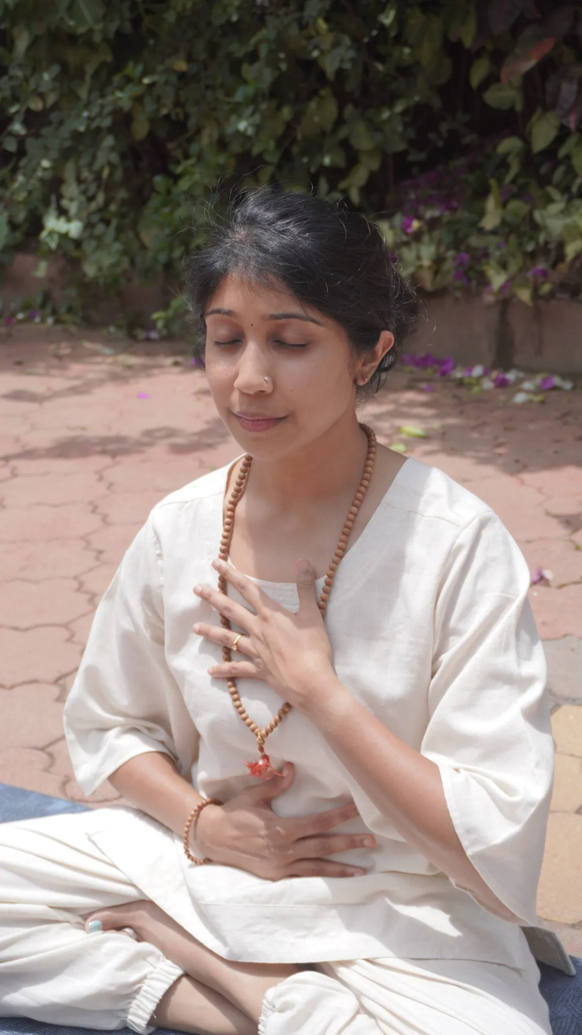 Jagruthi seated outdoors in a sunlit garden, one hand resting on her heart in quiet breath awareness