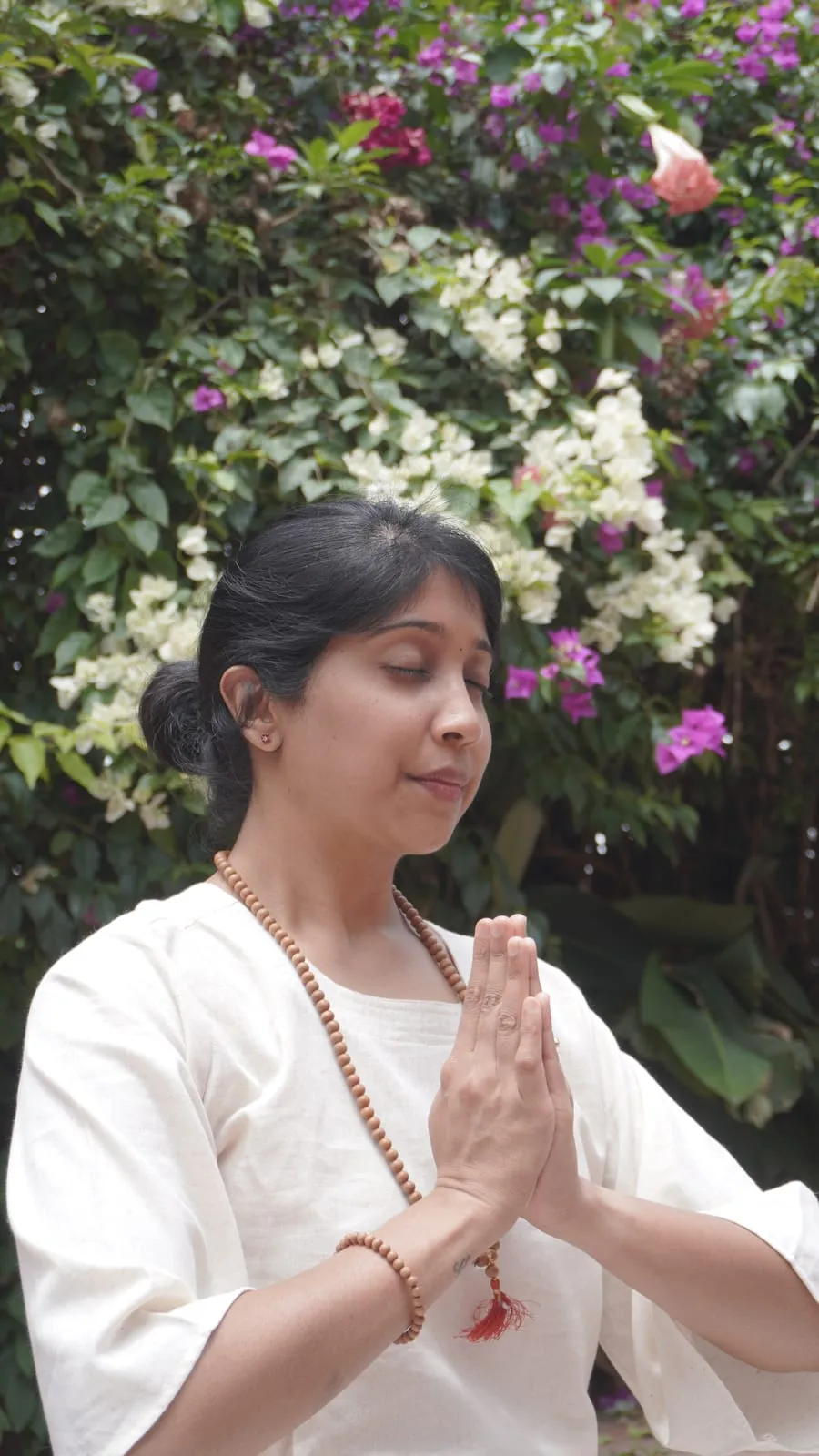 Jagruthi standing with her palms together at her chest and her eyes closed, in front of a wall of flowering bougainvillea