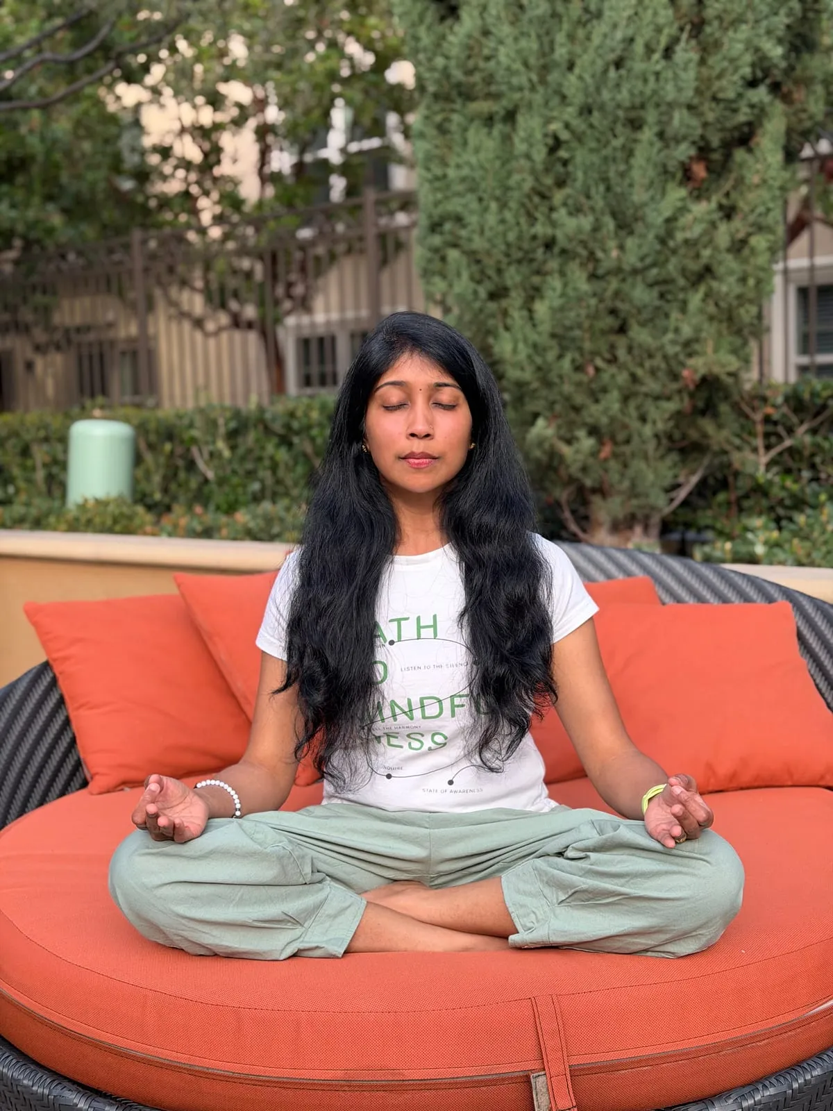 Jagruthi seated cross-legged with her eyes closed and her hands resting on her knees, meditating on a garden daybed