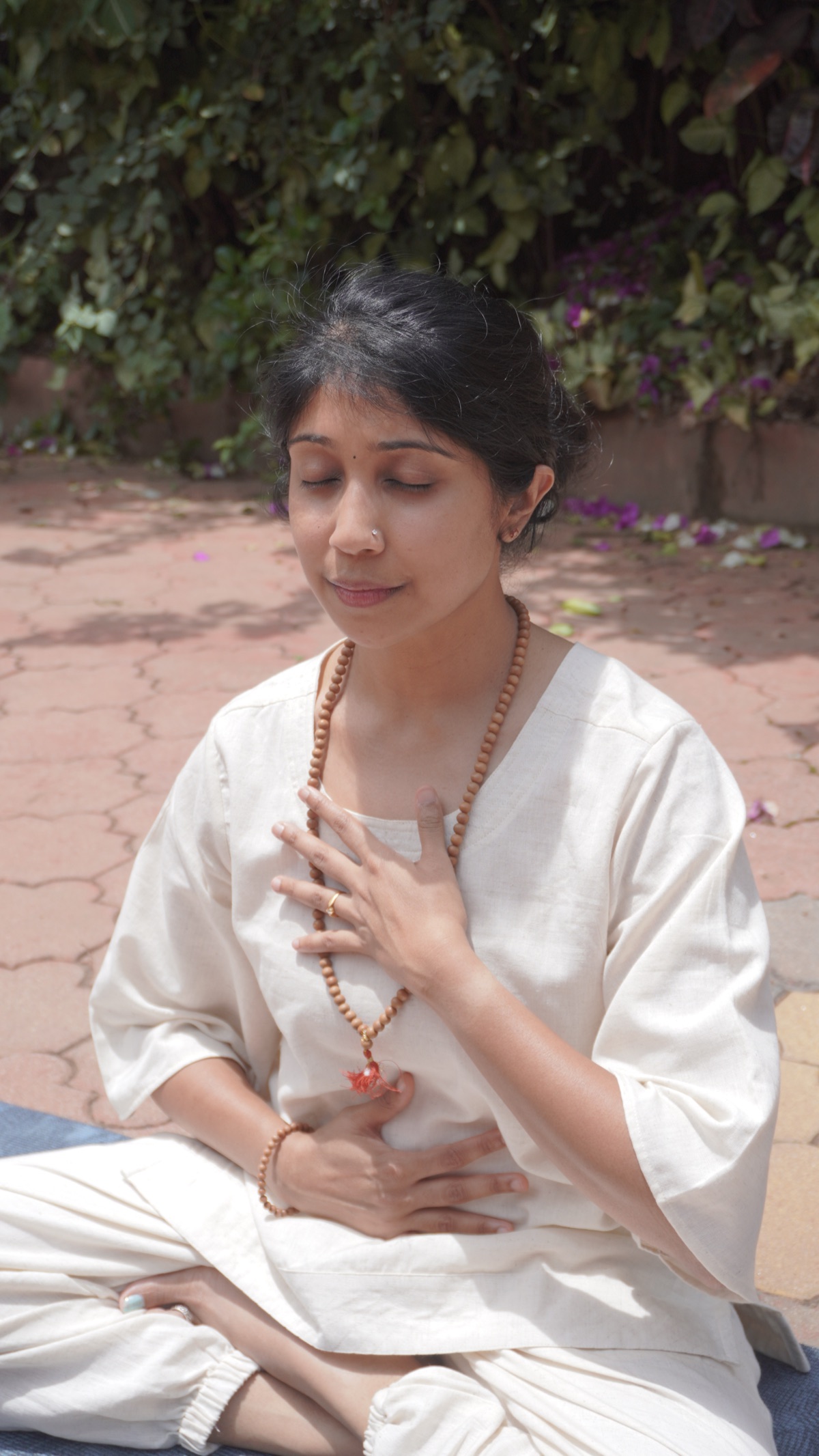 Jagruthi seated outdoors in a sunlit garden, one hand resting on her heart in quiet breath awareness