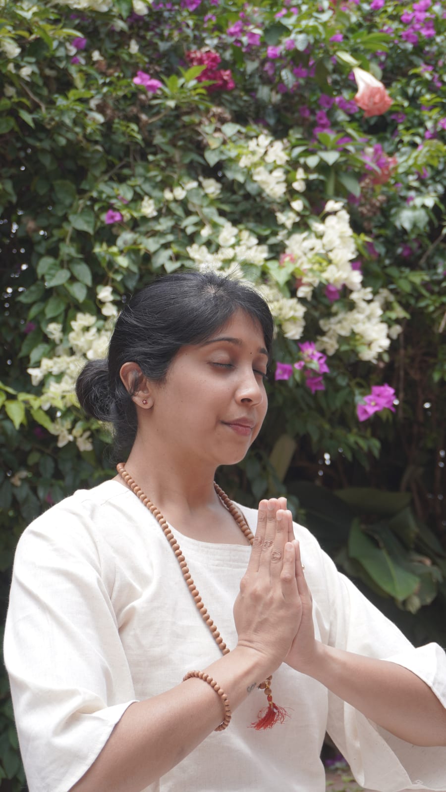 Jagruthi standing with her palms together at her chest and her eyes closed, in front of a wall of flowering bougainvillea