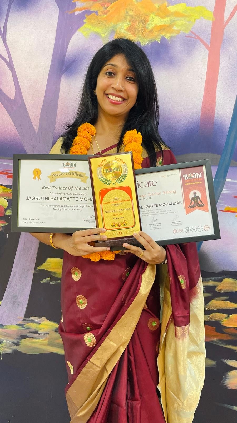 Jagruthi standing in a maroon sari and marigold garland, holding her Best Trainer of the Batch trophy and two framed yoga teacher training certificates
