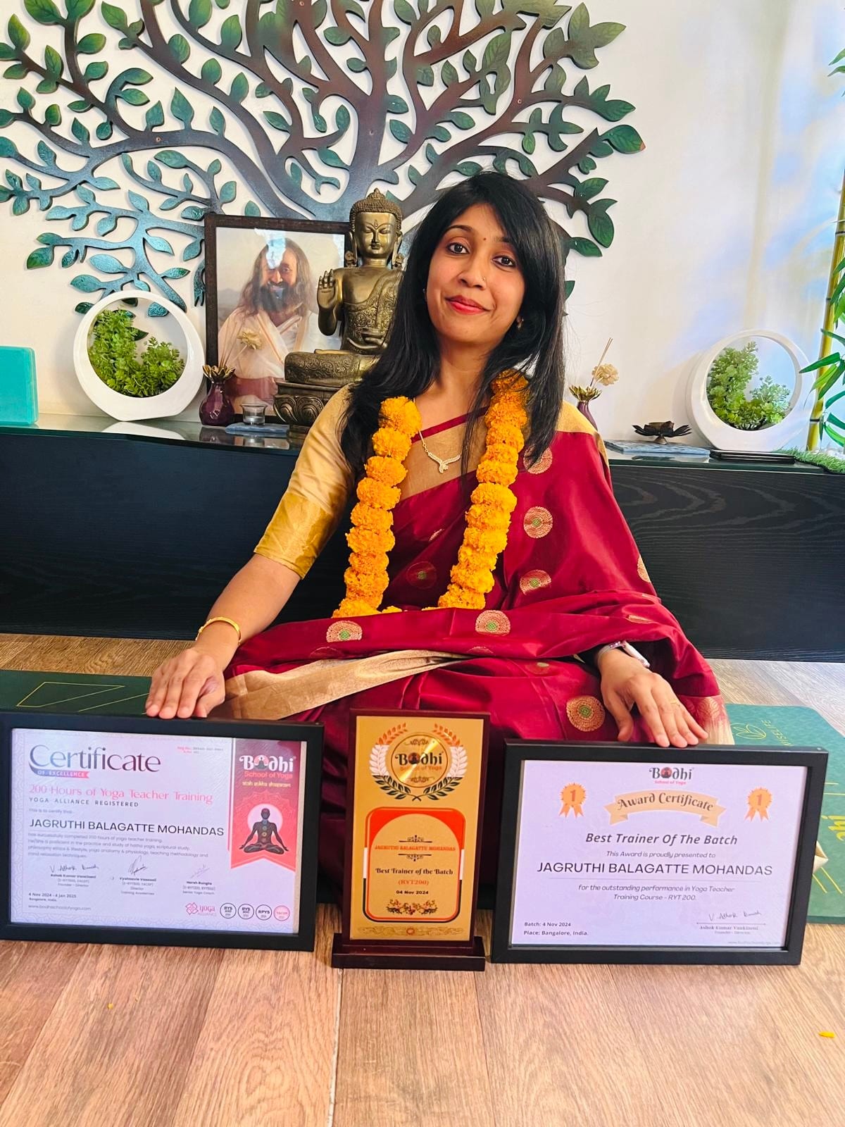 Jagruthi seated in a garland behind her Yoga Alliance certificate, trophy and Best Trainer award set out on the floor in front of her