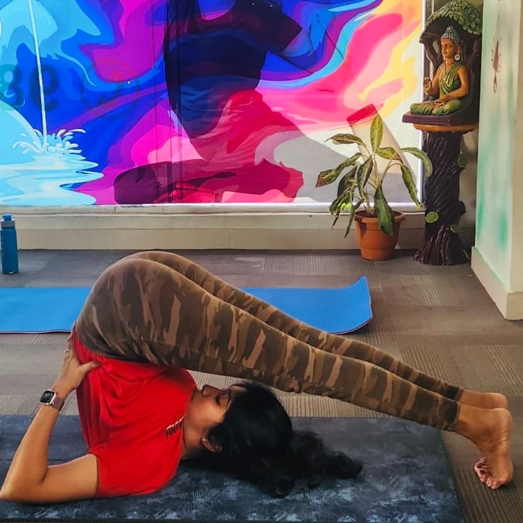 Jagruthi in halasana, the plough pose, lying on her back on a mat with her legs extended over her head to the floor