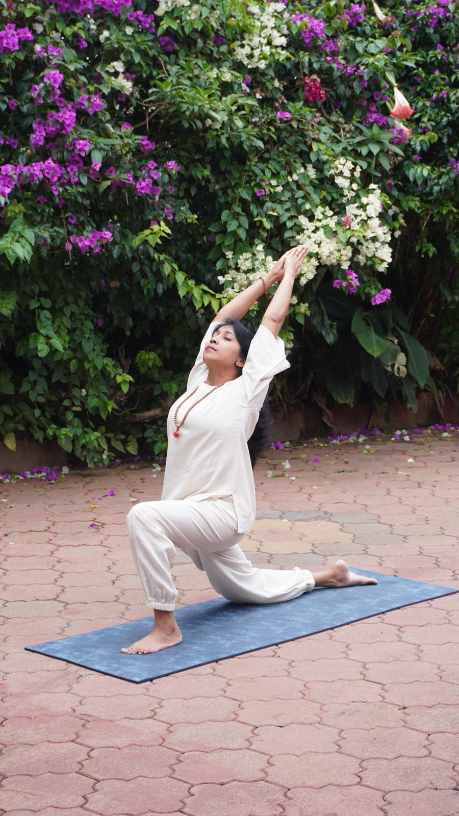 Jagruthi in a low lunge on a mat outdoors, both arms stretched overhead and her head tilted back