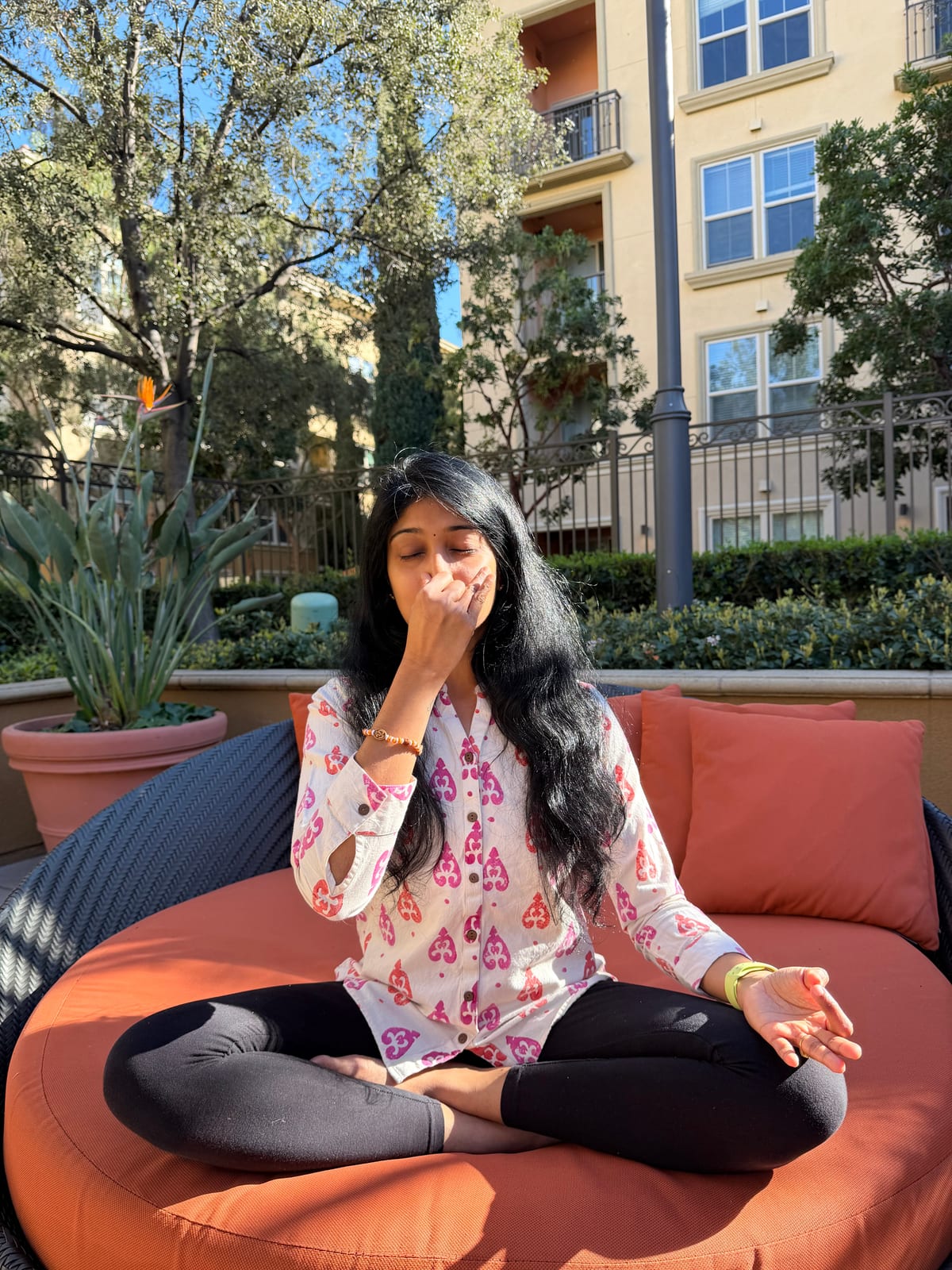 Jagruthi seated cross-legged on a round garden daybed, practising alternate-nostril breathing with her right hand at her nose and her left hand resting on her knee
