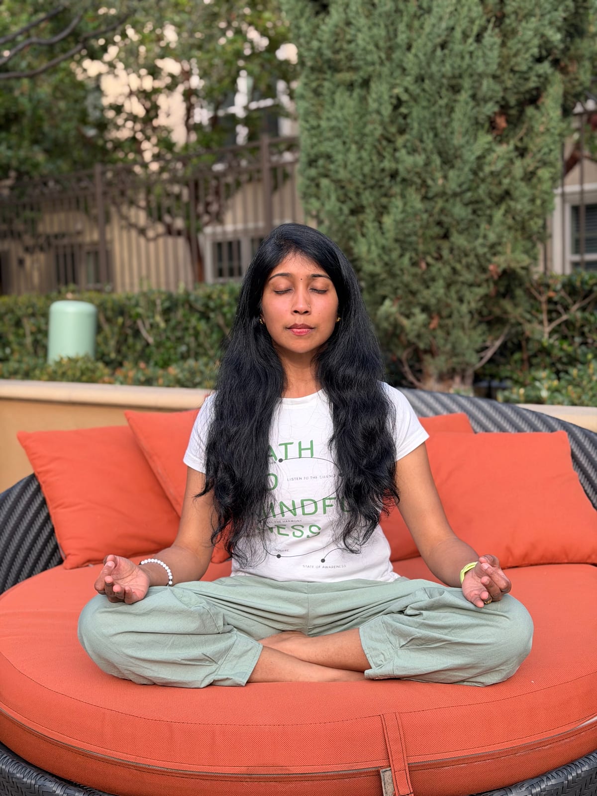 Jagruthi seated cross-legged with her eyes closed and her hands resting on her knees, meditating on a garden daybed